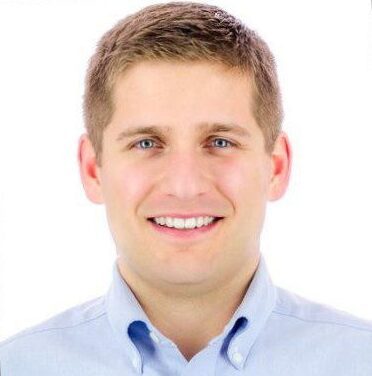 A man with short, light brown hair smiles while wearing a light blue button-up shirt. The background is plain white.